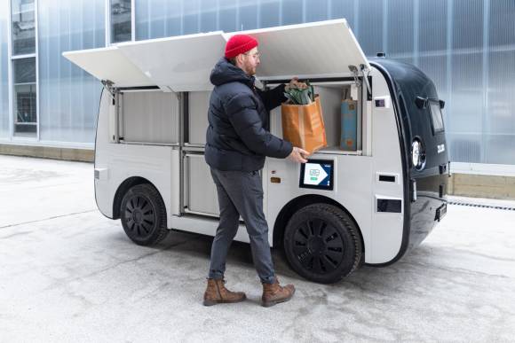 Ein Mann entnimmt einem der Ladefächer des autonomen Fahrzeuges einen gefüllten Migros-Papiersack 