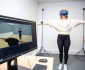 Eine Frau mit VR-Brille imitiert die Bewegung eines Avatars, das auf einem Bildschirm zu sehen ist